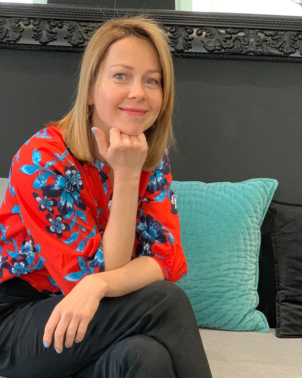 Elena Padurariu, smiling, sits on a grey sofa beside a turquoise cushion, her chin resting on her hand
